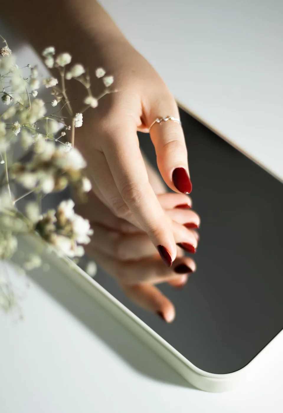 Glossy red manicure reflected in a mirror beside white flowers