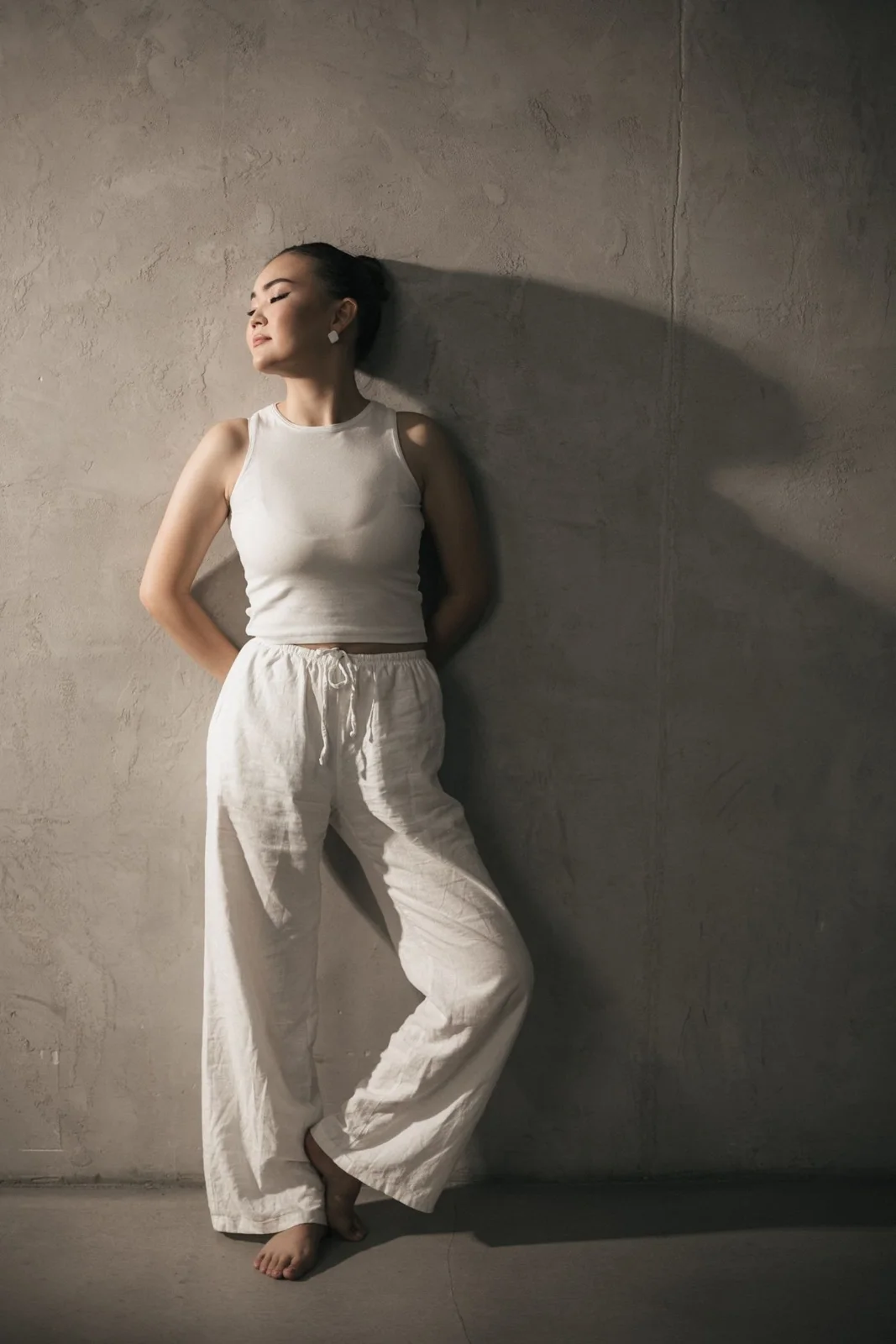 White sleeveless top with relaxed pale trousers against a concrete wall